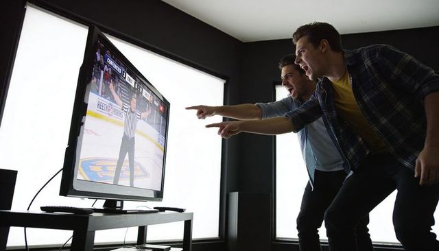 Two friends leaning forward cheering and pointing at live hockey game on living room tv