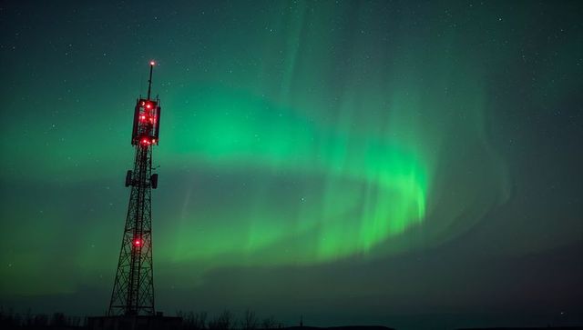 Emerald aurora curtain illuminating starry night over rural horizon with red-lit antenna tower
