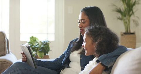 Denim-jacketed mom and daughter cuddling on sofa while sharing tablet in sunlit room