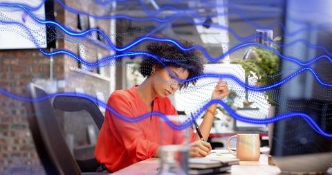 Focused woman writing at desk with blue data visualization overlays, notetaking, office