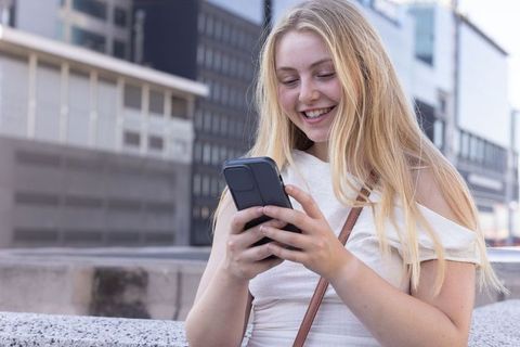 Smiling young woman texting on smartphone in urban plaza with modern architecture