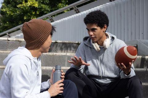 Diverse teen friends sitting on concrete steps holding american football and water bottle