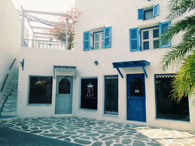 This image showcases a charming Mediterranean style courtyard with bright white walls contrasting with blue shutters and doors. Staircase with handrail is on the left, ascending to a small veranda. Elements of cozy and traditional Mediterranean architecture are present, including an ornate window cover and a potted tree. Ideal for use in travel brochures, architecture websites, real estate marketing, or decoration themes showcasing Mediterranean aesthetic.