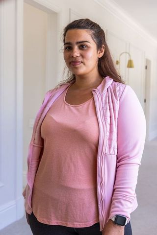 Indian woman standing in bright hallway wearing light pink quilted hoodie and smartwatch
