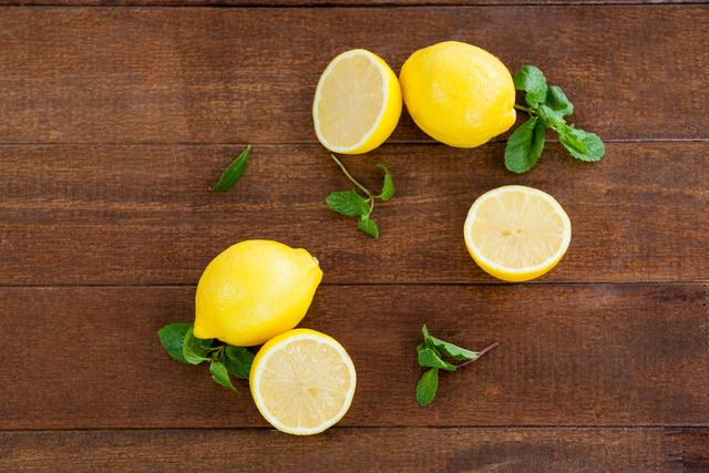 This image features fresh full and halved lemons along with mint leaves arranged on a wooden table. Ideal for use in food blogs, recipe books, health and wellness websites, and kitchen decor promotions. The vibrant colors and natural setting emphasize freshness and healthy living.