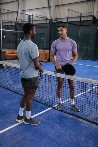 Two diverse male players chatting across net on indoor padel court holding round paddles