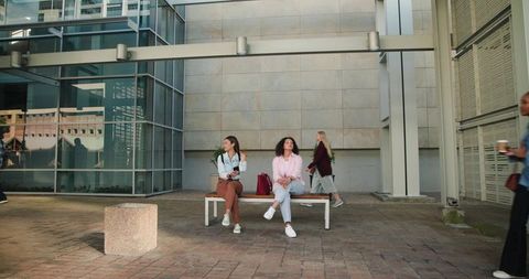 Young women sitting on bench in modern urban plaza holding smartphones and handbags on urban commute