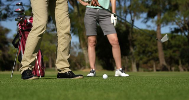 Two people stand on a sunny golf course, concentrating on a golf ball in the grass. Ideal for content related to sports, leisure activities, outdoor recreation, golf clubs, and athletic lifestyles.