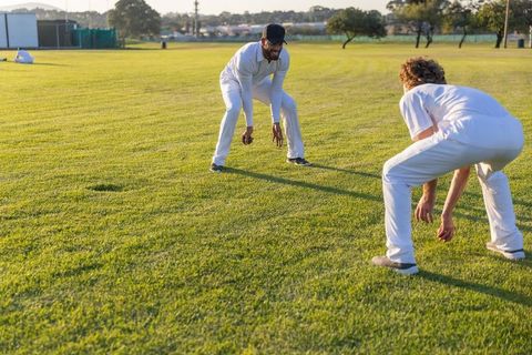 Two cricketers practicing close-in fielding on sunlit outfield during golden hour training