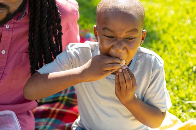 African American boy enjoying a snack while sitting next to his father on a sunny day. Ideal for use in family-oriented content, lifestyle blogs, advertisements promoting outdoor activities, and parenting articles. Highlights themes of bonding, happiness, and leisure.