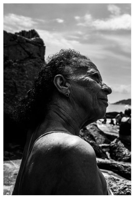 Elderly woman savoring the moment outdoors at a beach, featured in black and white. The image captures her in a peaceful and reflective state, standing by coastal rocks with the sea visible in the background. Perfect for themes related to aging, mindfulness, natural beauty, and outdoor lifestyles.
