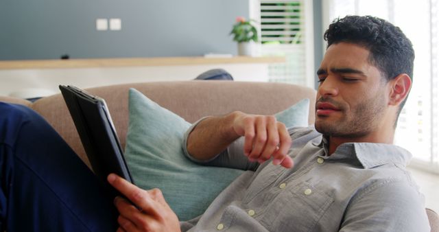 Young man resting on sofa at home while using tablet. Ideal for concepts related to home relaxation, modern lifestyles, technology usage, and indoor activities.