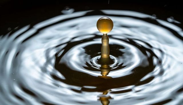Amber droplet balancing atop liquid column creating ripples with silvery reflection