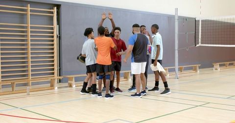 Diverse male volleyball team huddling and celebrating by raising hands in school gymnasium