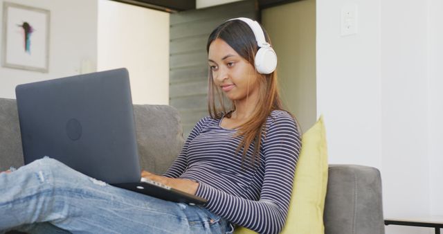 Image showcases woman relaxing comfortably on sofa while wearing headphones and using laptop. Perfect for concepts related to remote work, online learning, leisure, technology use, and lifestyle. Can be used for blog posts, articles, and advertisements focusing on home comforts, productivity, and digital lifestyles.