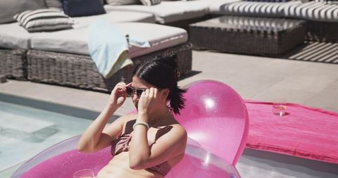 Woman adjusting sunglasses on pink inflatable float beside luxury poolside loungers