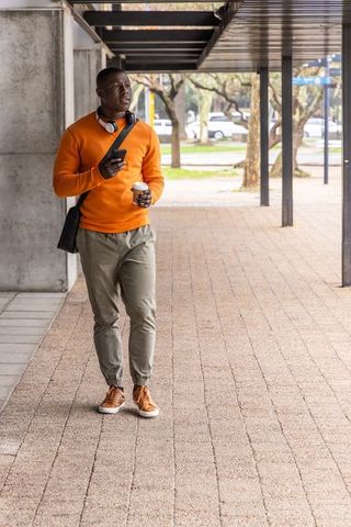 African american man walking with coffee and smartphone in orange sweater on urban campus