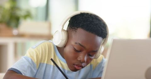 African-american student wearing headphones studying laptop writing in notebook, focused