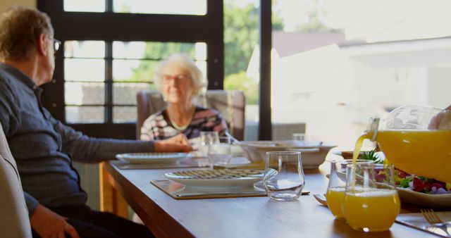 Elderly couple enjoying breakfast together at sunlit dining table, with focus on orange juice and plates. Ideal for concepts of senior lifestyle, retirement, morning routines, healthy eating, and family bonding. Can be used in promotions for retirement communities, health-related content, and family-oriented advertisements.