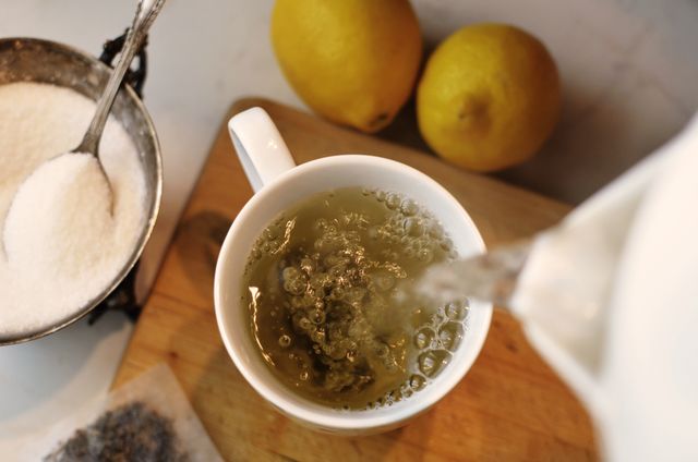This scene shows pouring of herbal tea from kettle into white cup on wooden board, surrounded by ingredients like lemons and sugar. Perfect for use in cooking blogs, cafes, restaurant menus, or marketing materials for tea and natural drinks retailers.