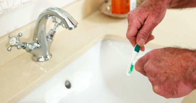 Photo shows an elderly man putting toothpaste on a toothbrush in a contemporary bathroom. The close-up image focuses on his hand over a sink and the faucet. This image can be used for promoting dental hygiene products, health care services and bathroom amenities.