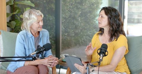 Two mature women recording conversational podcast in sunlit living room with microphones