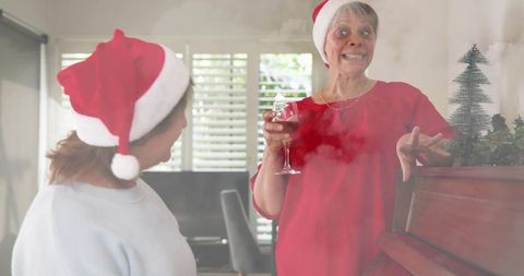 Senior woman toasting with wine and chatting by piano wearing santa hat in cozy festive home