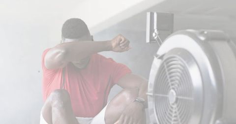 Tired athlete sitting on gym floor wiping sweat after intense indoor rowing session with fan