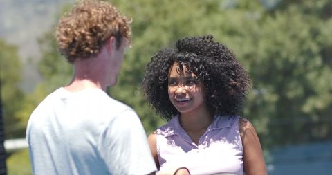 Sunlit multicultural couple smiling and talking on blue outdoor court in summer
