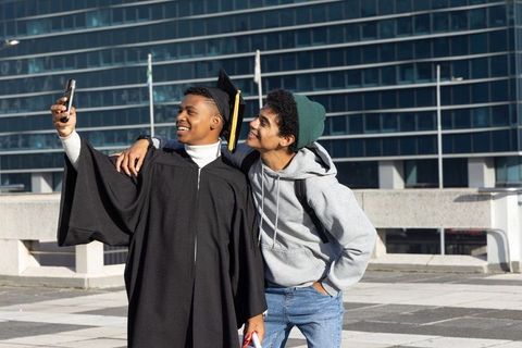 Diverse friends celebrating graduation taking selfie on urban terrace with cap, gown, diploma