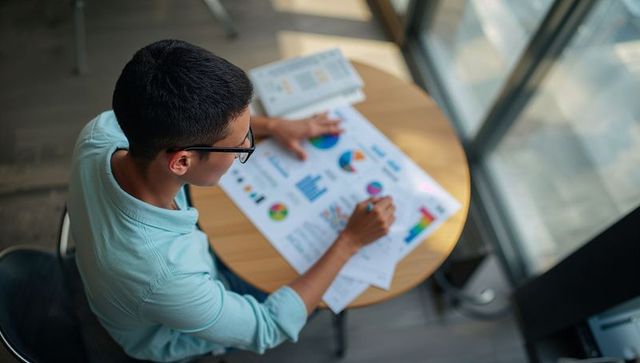 Student analyzing colorful charts and marking reports at wooden cafe table, overhead view