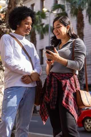 Multicultural friends sharing smartphone on sunny downtown street with crossbody bags