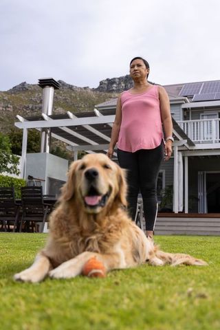 Mature woman standing and watching golden retriever with ball in solar-paneled backyard