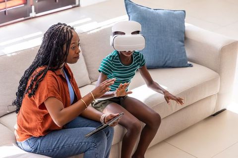 Black mother guiding daughter with vr headset while holding tablet on sunlit living room sofa