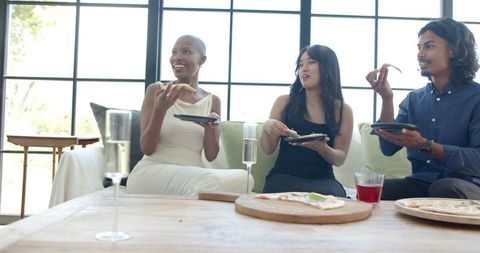 Friends enjoying pizza and chatting on sofa in bright modern living room with drinks