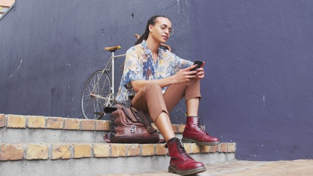 This video depicts a man with long dreadlocks in a casual outfit, sitting on stairs and smiling while using his smartphone. His vintage bicycle is leaning against the wall next to him. The sunny day and vibrant surroundings give a lively urban vibe. Ideal for use in lifestyle blogs, tech promotions, city living articles, or advertisements targeting modern, urban audiences.