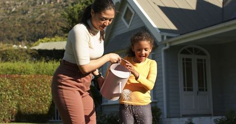 Mother and daughter playing with watering can in backyard, parenting bond and outdoor joy