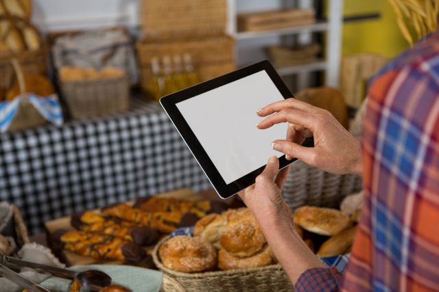 Female staff member using digital tablet at bakery counter, surrounded by various breads and pastries. Ideal for illustrating modern technology in small businesses, customer service in the food industry, or digital management in retail settings.