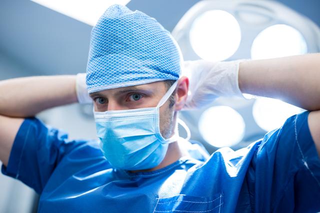 Surgeon wearing protective mask and scrub cap, adjusting gloves in well-lit operating room, indicating readiness for surgery. Useful for medical and healthcare topics, illustrating professional surgeons, hospital environment, and surgical preparation.