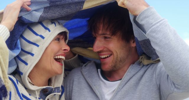 A young Caucasian couple shares a joyful moment under a blanket, with copy space. Their laughter and close proximity under the cozy cover suggest a romantic and playful relationship.