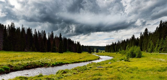 Scenic view of a peaceful mountain stream flowing through a lush green valley surrounded by dense forest under a dramatic sky with dark clouds. Perfect for nature enthusiasts, environmental campaigns, travel brochures, outdoor adventure promotions, and meditation or relaxation materials.