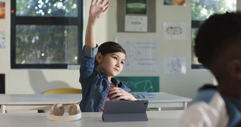 Asian girl raising hand in modern classroom using tablet and headphones