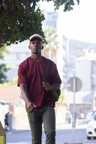 African american man walking under leafy canopy on urban sidewalk wearing cap and backpack