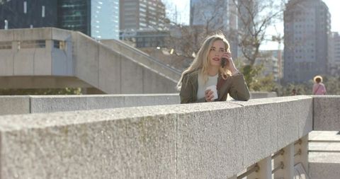 Young woman talking on smartphone while holding coffee on urban concrete bridge