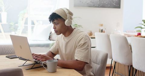African american man working remotely at sunlit dining table wearing headphones and beanie