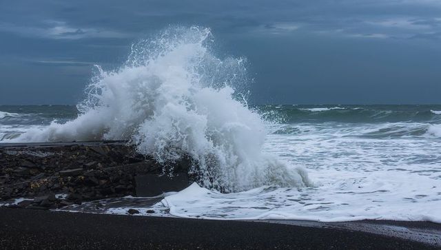 Storm waves crashing over breakwater on dark sand beach with exploding white foam