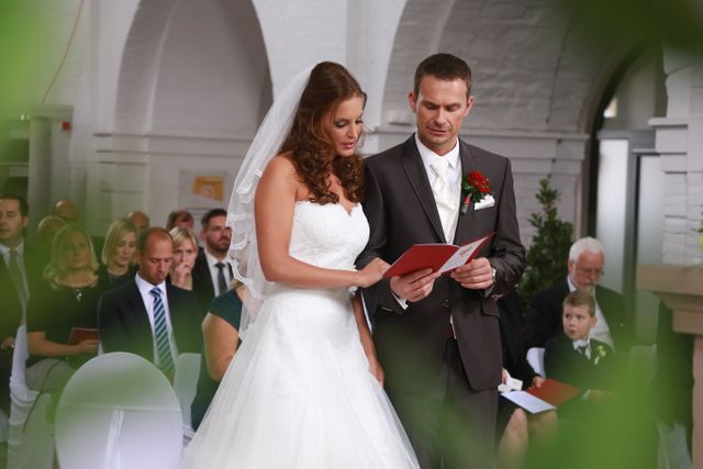 This image depicts a bride and groom exchanging vows during a wedding ceremony. The bride is wearing a beautiful white wedding dress, and the groom is in a formal suit with a red boutonniere. Guests are seated and watching the ceremony attentively. Ideal for use in wedding planning websites, marriage blogs, bridal magazines, and event planning services.