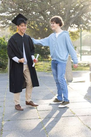 Sunlit campus graduate celebrating with friend, asian male in cap and gown holding diploma
