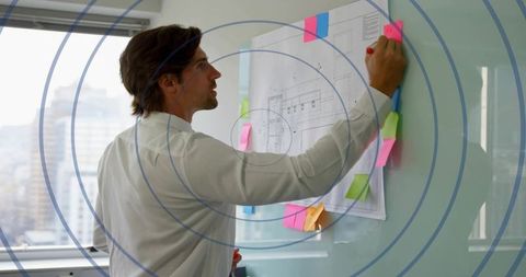 Man placing pink sticky note on glass board with architectural blueprint in modern office