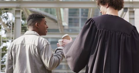 Colleagues sharing coffee while walking through modern glass atrium with robed professional